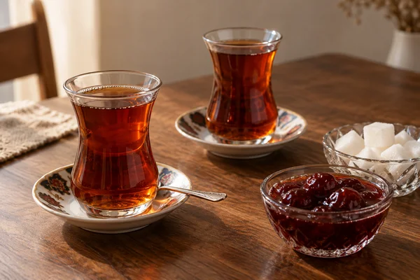 Two armudu glasses of amber tea on saucers beside sugar cubes and cherry preserve on a wooden table