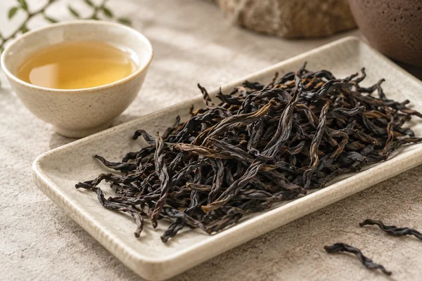 Long dark Dancong strips beside a pale gold tasting cup
