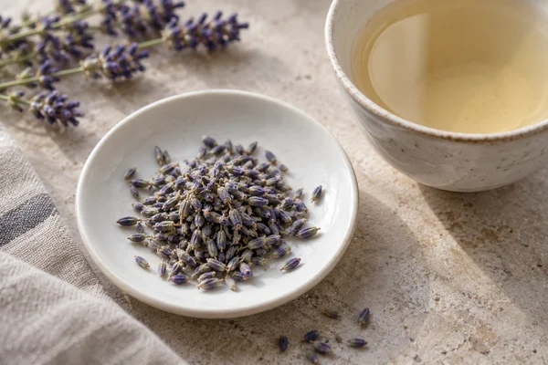 A small dish of dried lavender flowers beside a pale herbal infusion