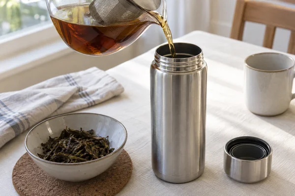 Filtered tea entering an open stainless steel flask beside drained tea leaves