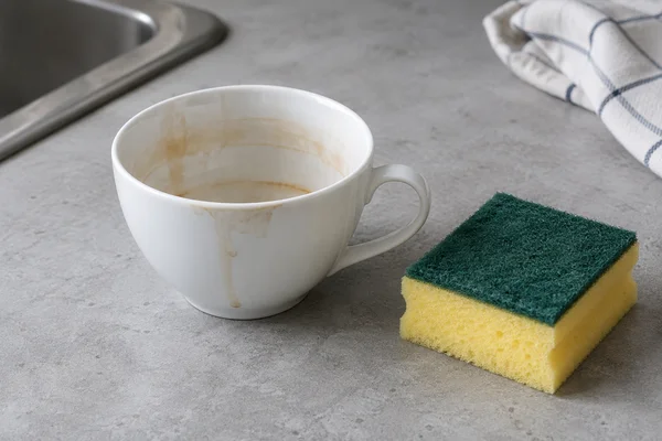 A cup with a visible tea stain beside a cleaning sponge